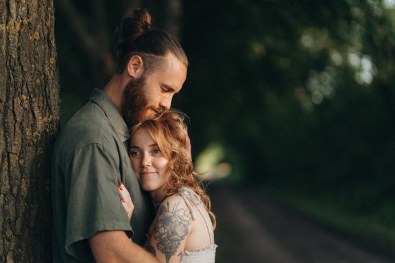 Braunhaariger Mann und rothaarige Frau umarmen sich, umgeben von Bäumen auf einem Weg.