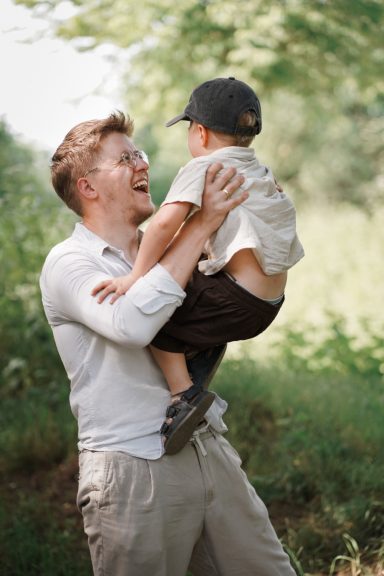 Vater hebt lächelnd seinen Sohn in der Natur hoch, umgeben von Bäumen.