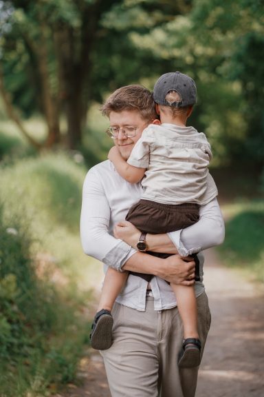Ein Vater trägt seinen kleinen Sohn auf dem Rücken, umgeben von grüner Natur.